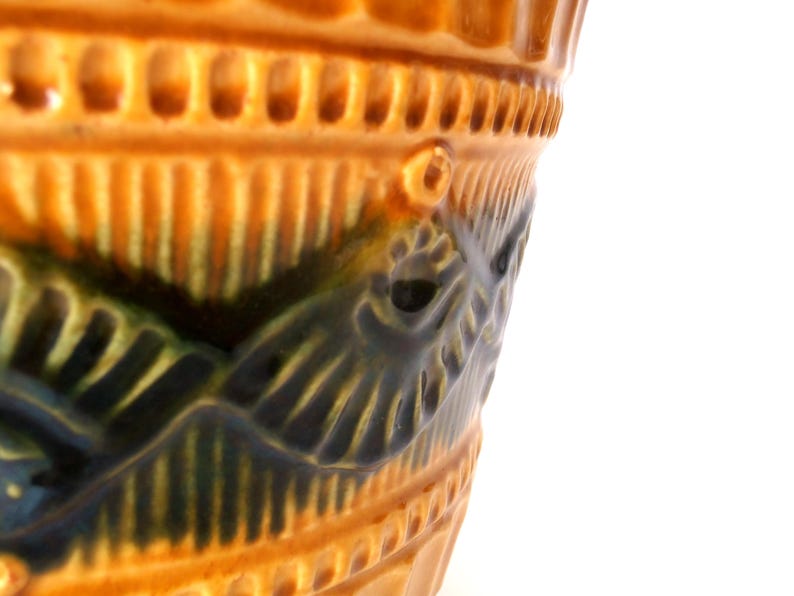 May include: A glazed ceramic vessel featuring a warm amber-orange base color with a prominent dark green, embossed wave-like pattern encircling the exterior. The rim displays a textured, repeating vertical notch design.