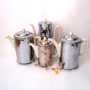 Könnte beinhalten: Vier silberne Kaffeekannen mit elfenbeinfarbenen Griffen und Deckeln. Die Kannen stehen auf einer weißen Oberfläche mit kleinen Holzfiguren davor.