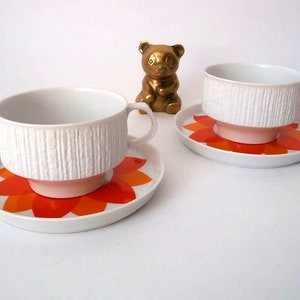 Puede incluir: Dos tazas de té blancas con lados texturizados y asas, colocadas sobre platillos a juego. Los platillos presentan un vibrante diseño floral naranja y rojo. Una pequeña figura de oso dorado se encuentra en el fondo.
