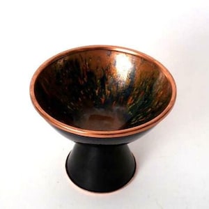 Puede incluir: Un cuenco con pedestal que presenta una amplia abertura circular con un borde de cobre metálico. El interior del cuenco muestra un acabado moteado y multicolor con tonos verdes, negros y naranjas. La base exterior es de color negro mate.