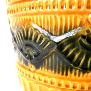 May include: A ceramic vessel featuring a vibrant orange glaze with a glossy finish. The surface is decorated with a raised, embossed pattern of dark green, fan-shaped motifs arranged in a repeating band around the exterior body.