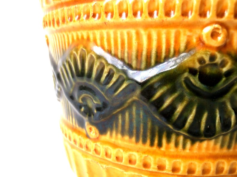 May include: A ceramic vessel featuring a vibrant orange glaze with a glossy finish. The surface is decorated with a raised, embossed pattern of dark green, fan-shaped motifs arranged in a repeating band around the exterior body.