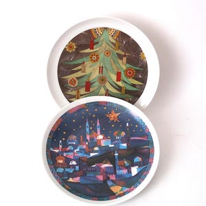 May include: Two white ceramic plates with colourful designs. One plate features a Christmas tree with candles, the other depicts a cityscape under a starry night sky. The plates have a white border.