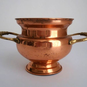 Könnte beinhalten: Ein Kupfertopf mit zwei Messinggriffen. Der Topf hat einen abgerundeten Boden und eine breite, runde Öffnung.