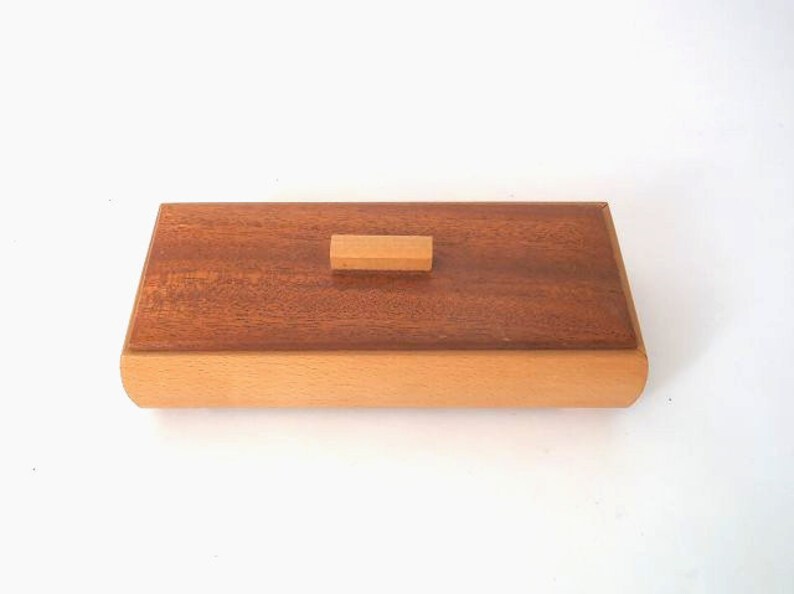 May include: A rectangular wooden box with a light brown lid and a dark brown base. The lid has a small rectangular wooden knob.