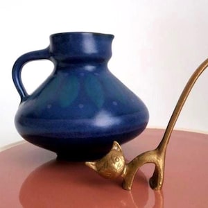 May include: A dark blue ceramic pitcher with a rounded body and a single handle, positioned next to a small, stylized brass cat figurine with an elongated tail and etched facial features, both set against a plain white background.