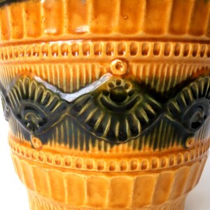 May include: A cylindrical ceramic vessel featuring a glossy mustard yellow glaze with vertical ribbing. A central horizontal band displays a dark green, relief-molded pattern of repeating fan-like shapes and small circular accents.