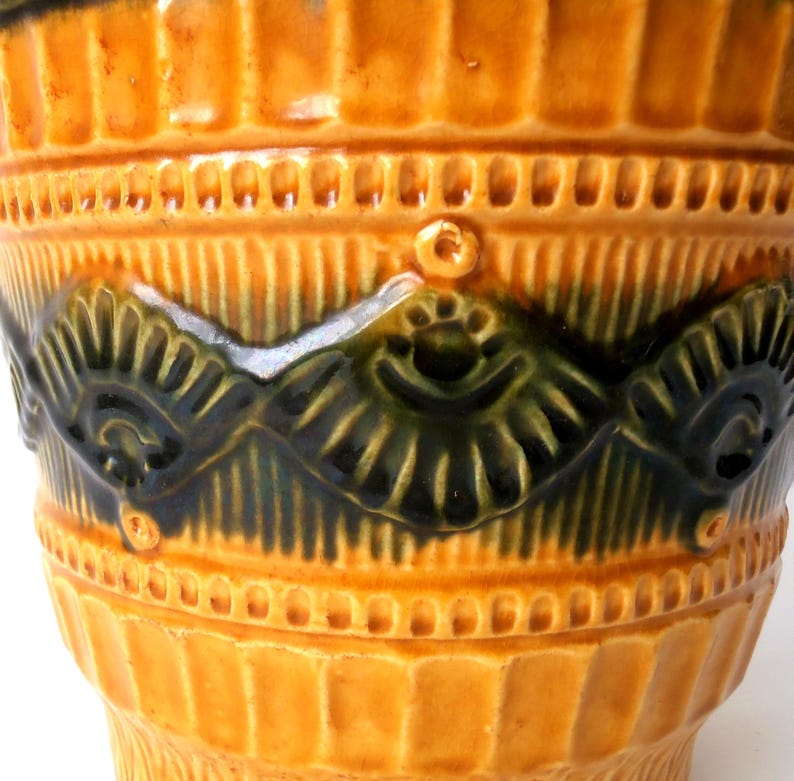 May include: A cylindrical ceramic vessel featuring a glossy mustard yellow glaze with vertical ribbing. A central horizontal band displays a dark green, relief-molded pattern of repeating fan-like shapes and small circular accents.