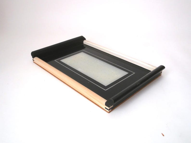 May include: A black tray with a wooden frame and a white border around the inside. The tray has rolled edges.