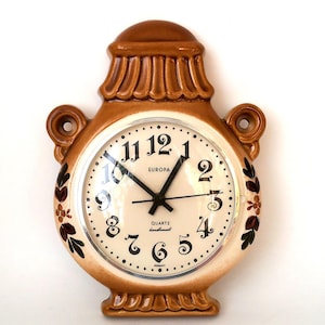May include: A brown ceramic wall clock with a cream-coloured face. The clock has black numerals, black hands, and the word "EUROPA" printed on the face. The clock is decorated with floral accents and the words "QUARTZ handmade GERMANY".