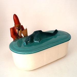 Può includere: Un piatto rettangolare in ceramica bianco sporco con coperchio turchese con un pesce scolpito. Una piccola statuina di legno con cappotto e cappello rossi è vicina. Il piatto è probabilmente per servire o conservare.