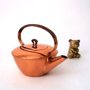 May include: A polished copper teapot with a rounded body, a short spout, and a lid featuring a small handle. The arched top handle is wrapped in dark brown material. Beside the teapot sits a small, seated brass bear figurine with dark eyes.