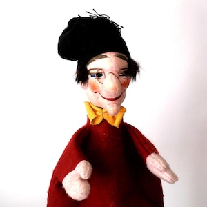 May include: A hand puppet featuring a figure with a pale face, rosy cheeks, and dark hair wearing a black cap and round wire-rimmed glasses. The figure has a yellow collar and a solid red body with light-colored hands extending from the sleeves.