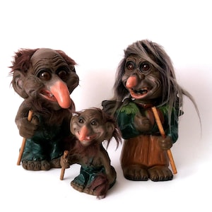 May include: Three troll figurines with exaggerated features. The trolls have large noses, wide smiles, and are dressed in green and brown clothing. Each troll holds a wooden stick. The figurines are set against a white background.