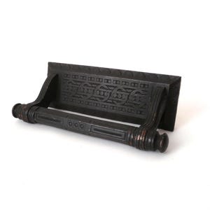 Puede incluir: Toallero de metal negro con un diseño ornamentado. La barra tiene una base rectangular con un patrón decorativo y dos brazos curvos.