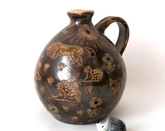 May include: A brown ceramic pitcher with a handle, decorated with a repeating pattern of stylized hedgehogs and flowers. The pitcher is sitting on a white surface, with a small ceramic hedgehog figurine in front of it.