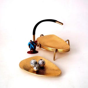 May include: A gold-colored, hammered metal serving tray with three legs and a curved handle. A smaller, matching tray is in front, holding three decorative spheres. A Superman figurine is next to the tray.