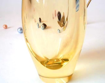 May include: A tall, amber-colored glass pitcher with a smooth, rounded body and a thin, curved handle extending from the side. The translucent glass reflects light and surrounding objects, creating a warm, golden hue throughout the vessel.