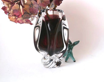 May include: A dark purple glass vase with a unique, abstract design. The vase has a twisted, organic shape with a wide opening at the top and a narrow base.