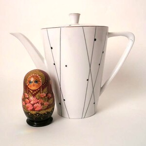 Op de afbeelding: Een witte theepot van keramiek met een zwart-wit geometrisch ontwerp. De theepot heeft een tuit en een handvat. Een kleine, houten, beschilderde pop staat op de voorgrond.