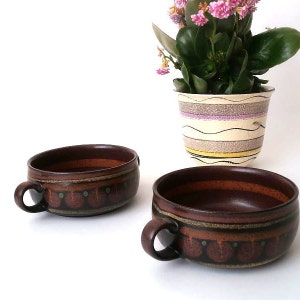 May include: Two brown ceramic soup bowls with handles, featuring a decorative band of orange and green dots. A potted plant with pink flowers sits in the background. The bowls have a rustic, handmade appearance.