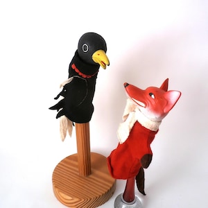 May include: Two vintage hand puppets: a black crow with a yellow beak and red trim, and a red fox with a white scarf. The crow is mounted on a wooden stand. The fox is on a glass and metal stand. Puppets are likely for children.