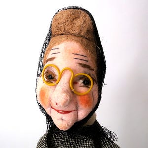 May include: A handcrafted doll head featuring light skin with rosy cheeks, a red painted mouth, and brown eyebrows. The doll wears round yellow plastic glasses and a black mesh head covering over a textured brown hair bun, with a black and white patterned collar.