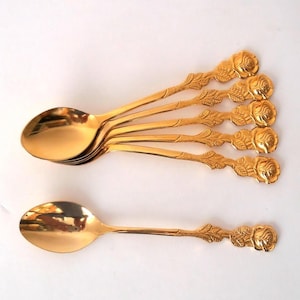 Puede incluir: Un conjunto de seis cucharas doradas. Cada cuchara tiene un cuenco redondeado y un mango con un diseño de rosa. Las cucharas están dispuestas sobre una superficie blanca, con una cuchara separada.