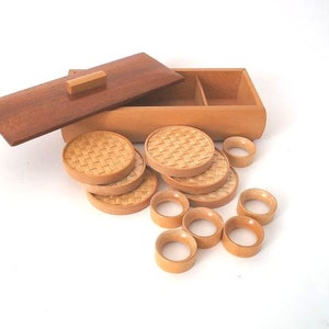 May include: A set of wooden coasters and napkin rings in a wooden box. The coasters are round and have a woven pattern. The napkin rings are also round and have a smooth finish.