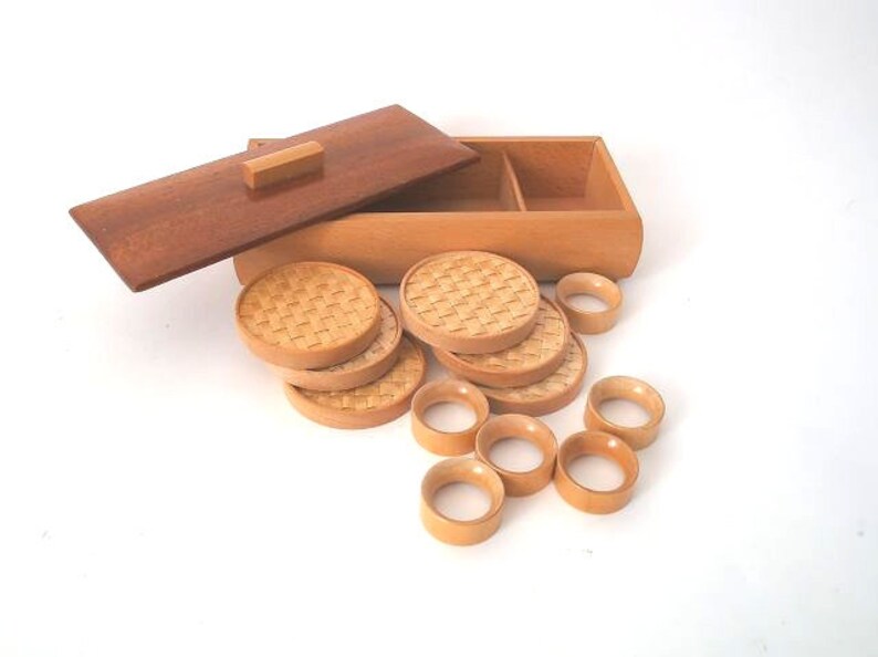 May include: A set of wooden coasters and napkin rings in a wooden box. The coasters are round and have a woven pattern. The napkin rings are also round and have a smooth finish.
