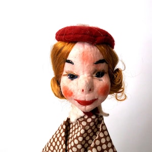 May include: A doll head with ginger hair and a red beret. The face features painted black eyebrows, dark eyeliner, rosy cheeks, and red lips. The doll wears a brown collared shirt with a white polka dot pattern and a white collar underneath.