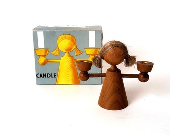 Puede incluir: Un portavelas de madera con forma de niña con dos coletas. El portavelas tiene dos brazos para velas y está hecho de madera marrón oscura. La caja dice "CANDLE" en ella.