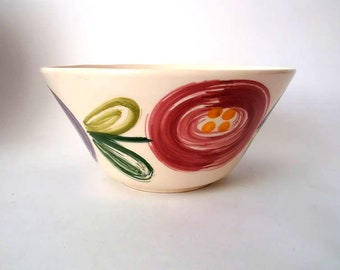 May include: A white ceramic bowl with a hand-painted design of pink, green, and purple flowers. The bowl has a wide rim and a rounded bottom.