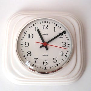 May include: A white square wall clock with a round face. The clock face is white with black numerals and hands, and a red second hand. The clock reads "EUROPA" and "QUARTZ". The clock has a silver bezel and is made in Germany.