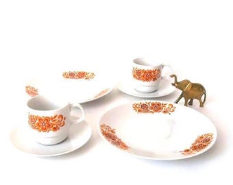 May include: A white ceramic dinnerware set featuring two cups with matching saucers and two oval plates, all decorated with an orange and brown floral pattern. A small brass elephant figurine stands next to the cups and plates on the white surface.