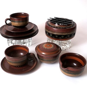 May include: A collection of brown ceramic tableware featuring two cups with matching saucers, a lidded bowl, a small creamer, and a circular warmer with a black metal grate. Each piece displays horizontal bands of orange, tan, and dark brown glaze.