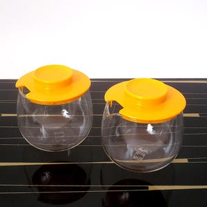 Puede incluir: Dos tarros de cristal transparente con tapas de color amarillo brillante. Los tarros tienen forma redonda y están sobre una superficie negra con rayas doradas. Las tapas tienen un diseño curvo. Los tarros parecen ser vintage.