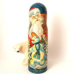 Può includere: Una matrioska cilindrica in legno con una scena dipinta a mano raffigurante Babbo Natale con la barba bianca e un pupazzo di neve con sciarpa verde e cappello arancione. Una piccola statuetta di orso polare color crema si trova dietro.