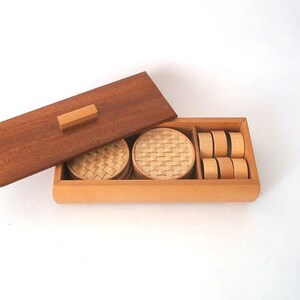 May include: A wooden box with a lid, containing woven coasters and round wooden coasters. The box is made of light wood and has a dark wood lid.