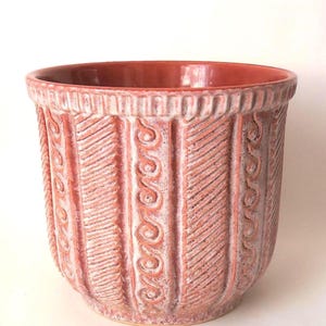 Peut inclure: Un pot de fleur en céramique de couleur terre cuite avec un motif sculpté texturé. Le pot présente des motifs verticaux nervurés et des volutes décoratives. L'intérieur et le bord sont d'une teinte plus foncée.
