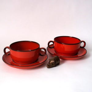 Peut inclure: Deux bols à soupe rouges avec anses et soucoupes assorties, bordés de noir. Une petite sculpture en bronze d'animal est placée entre les bols. Service de table classique.