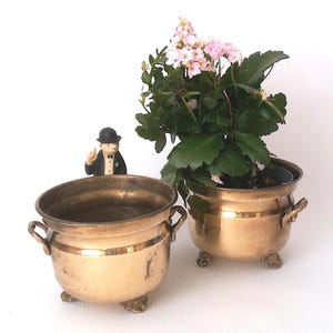 Op de afbeelding: Twee plantenbakken van gepolijst messing met ronde lichamen, kleine handvatten aan de zijkant en drie versierde voetjes. De ene is leeg, terwijl de andere een groene plant met kleine roze bloemetjes bevat. Een klein figuurtje in pak is zichtbaar.