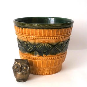 May include: A ceramic plant pot featuring a vibrant mustard yellow glaze with a dark green band around the top and a textured, repeating fan-like pattern. Beside the pot sits a small, metallic bronze-toned owl figurine with detailed feathers.