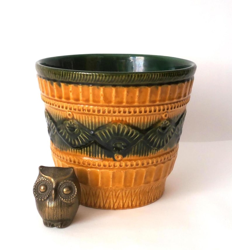May include: A ceramic plant pot featuring a vibrant mustard yellow glaze with a dark green band around the top and a textured, repeating fan-like pattern. Beside the pot sits a small, metallic bronze-toned owl figurine with detailed feathers.