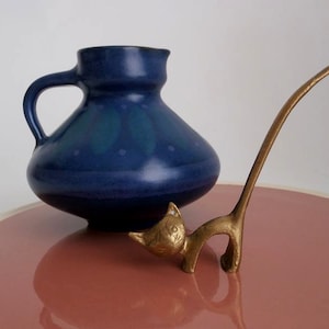May include: A blue ceramic pitcher with a white and blue design sits on a pink plate next to a gold metal cat figurine.