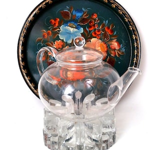 May include: A clear glass teapot with a lid and a handle sits on a clear glass stand. The teapot is on top of a black tray with a painted floral design.