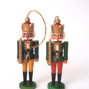 Op de afbeelding: Twee houten notenkraker-ornamenten op groene ronde voetstukken. De ene notenkraker draagt een blauw jasje met een gele broek, de andere een groen jasje met een rode broek. Beide hebben gouden kronen, wit haar en houden gouden staven vast.