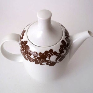 Puede incluir: Tetera blanca con un patrón floral marrón. La tetera tiene una tapa y un pico.