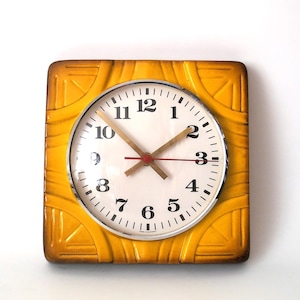 May include: A square wall clock with a mustard yellow ceramic frame featuring embossed geometric patterns. The clock face is white with black Arabic numerals, a silver-toned circular bezel, and three hands, including a thin red second hand.