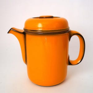 Può includere: Una teiera in ceramica color arancione brillante con finitura lucida e coperchio abbinato. La teiera presenta un corpo cilindrico, un manico curvo e un beccuccio corto, il tutto accentuato da sfumature marrone scuro lungo i bordi.
